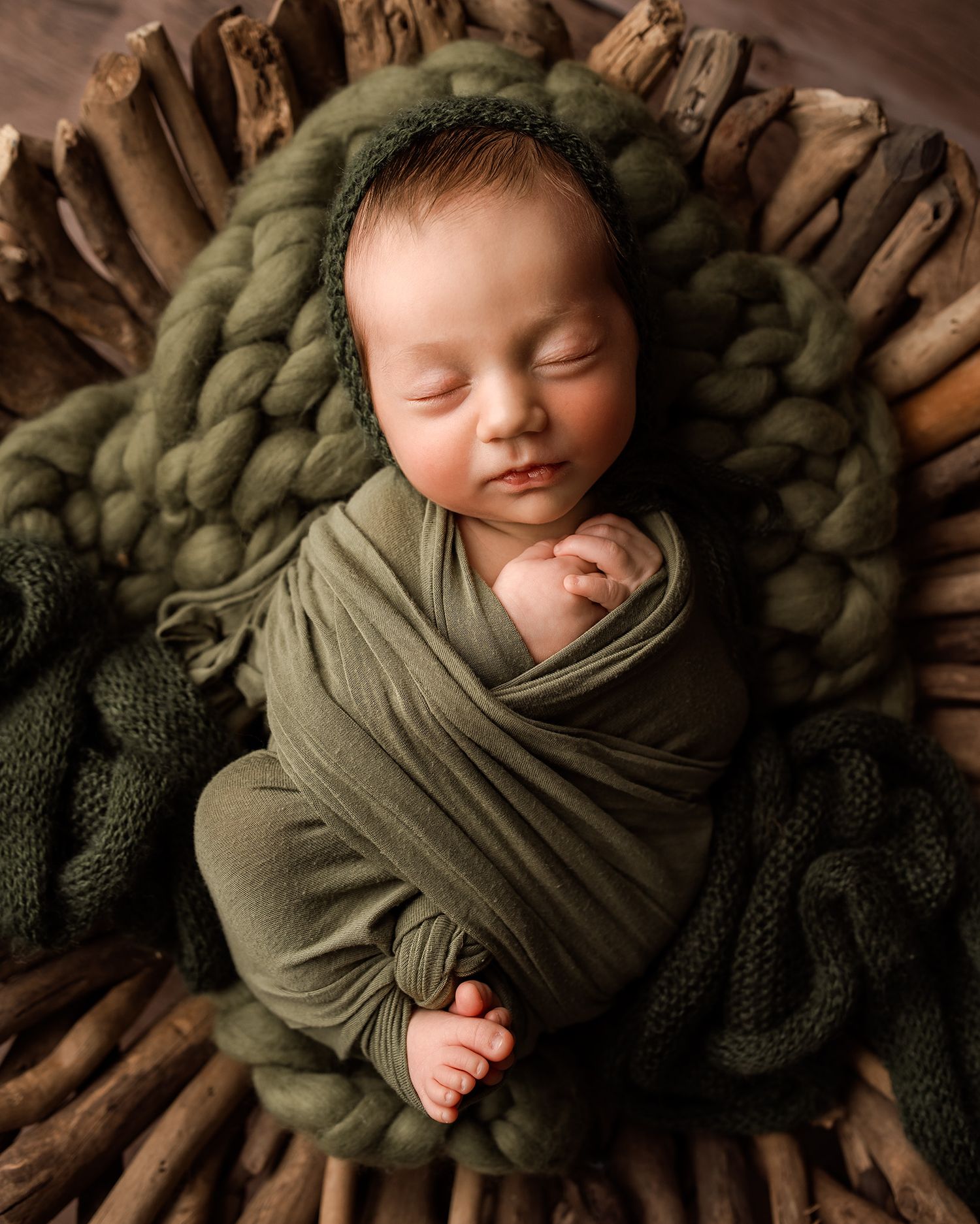 Newborn baby swaddled in olive green wrap sleeping peacefully in a rustic wooden bowl with chunky knit blanket.