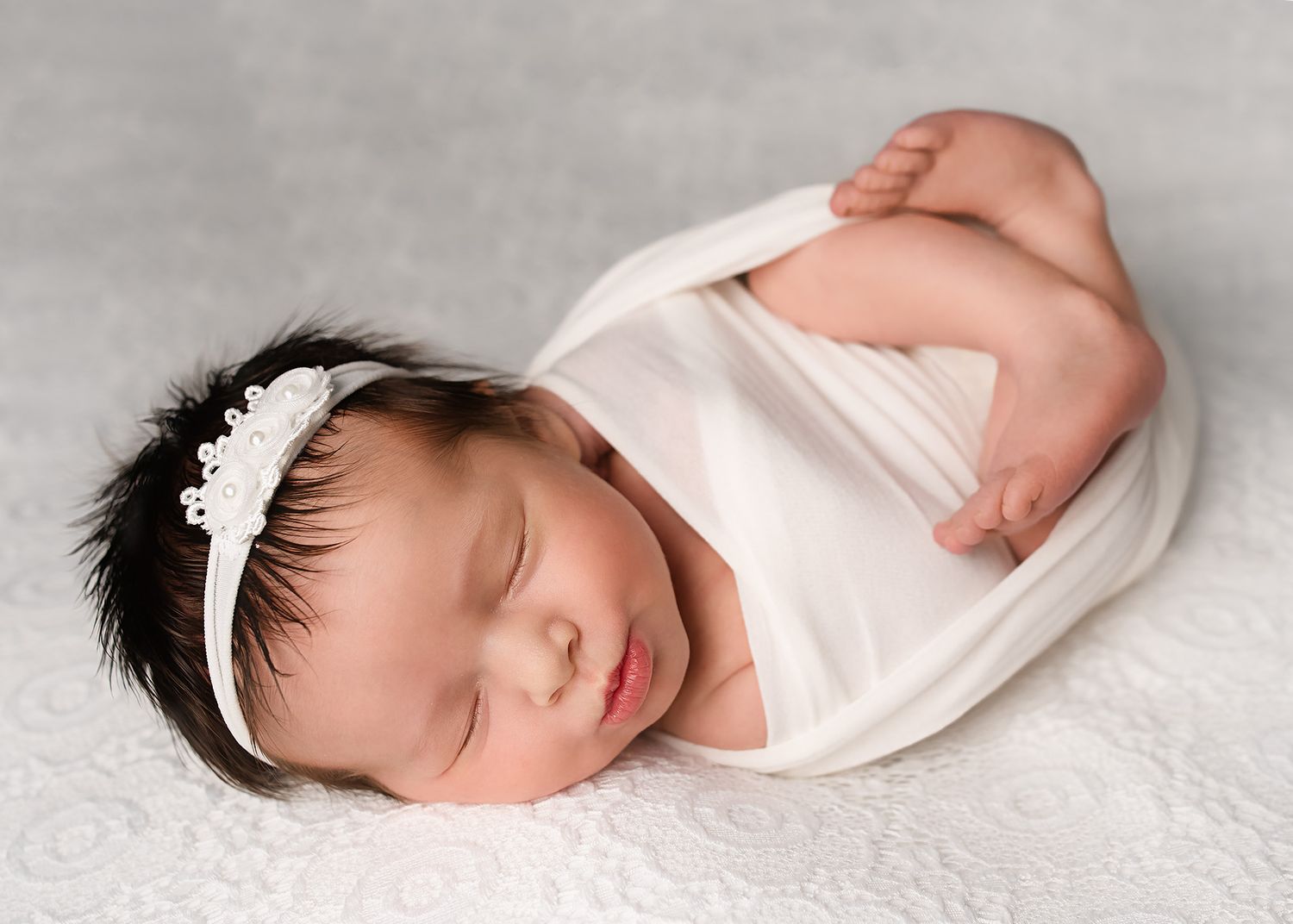 Sleeping newborn baby girl wrapped in white swaddle with lace headband on a soft white background.
