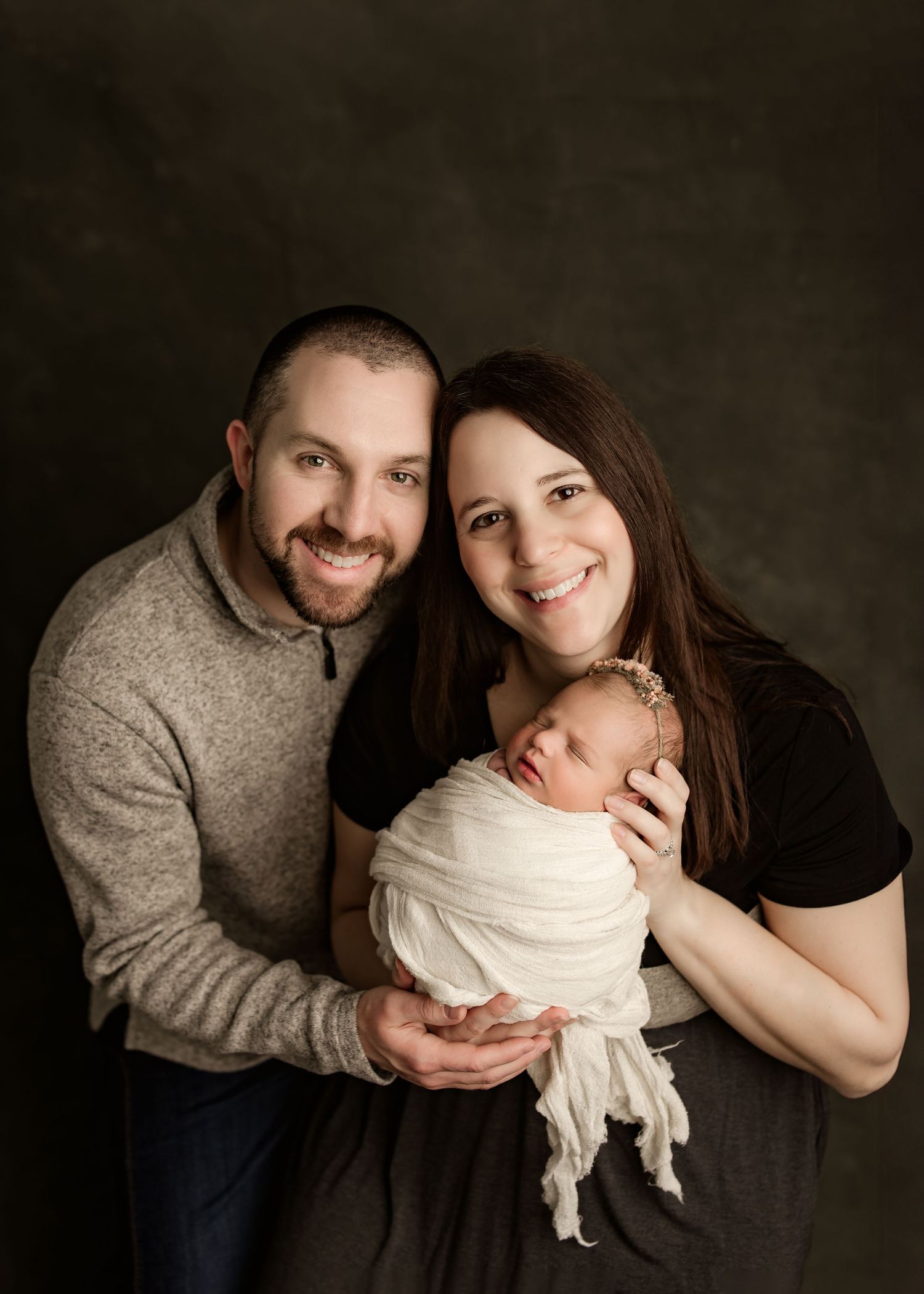 Smiling parents holding their swaddled newborn baby in a professional studio portrait photo.