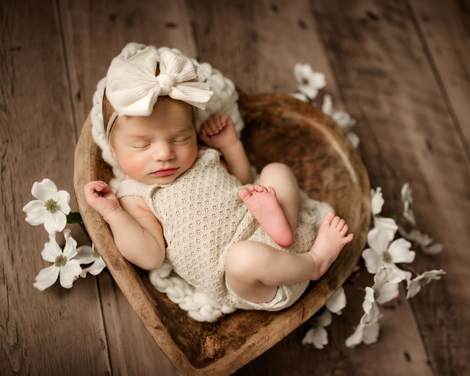 Newborn baby sleeping in heart-shaped wooden bowl wearing cream knit outfit and bow headband, surrounded by white flowers.