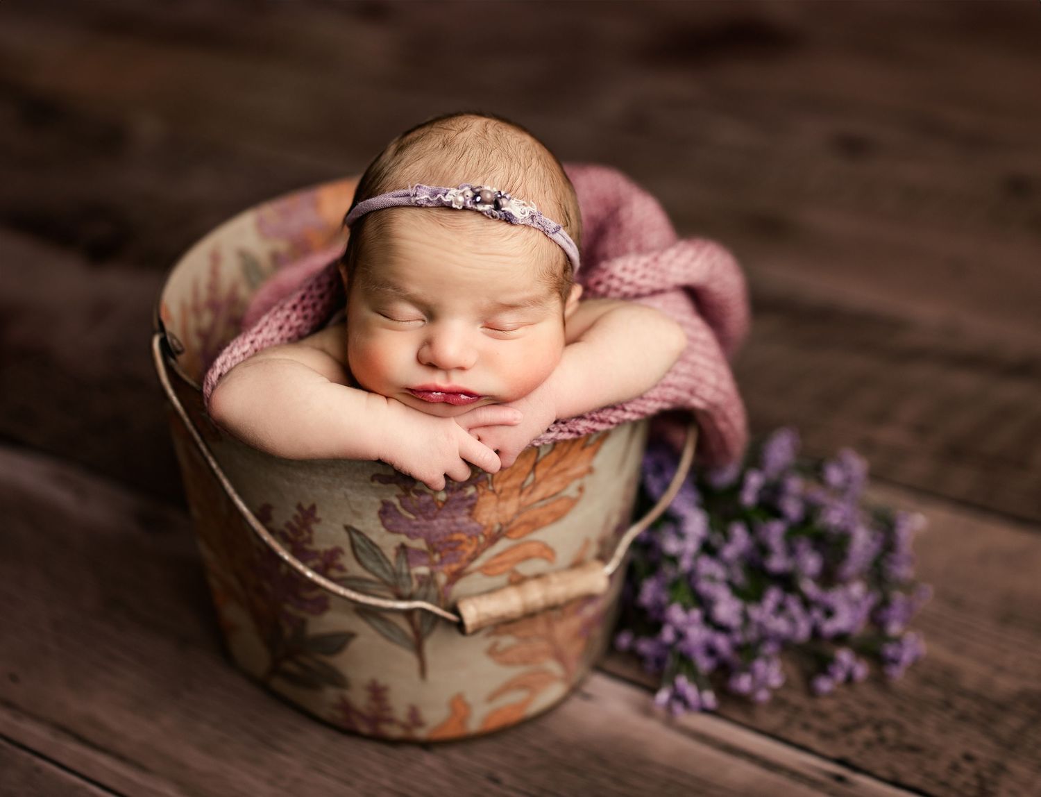Sleeping newborn baby girl in floral bucket with pink wrap and headband beside purple lavender flowers.