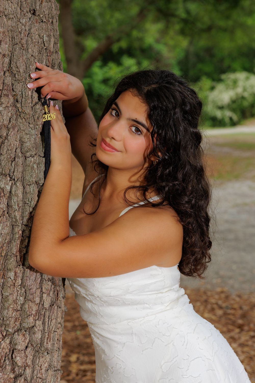 Young woman in white dress leaning against tree trunk in park, with curly dark hair and gold bracelet.