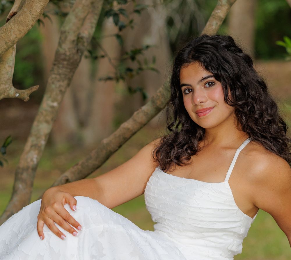 Young woman in white dress sitting gracefully under trees in a lush green outdoor setting.