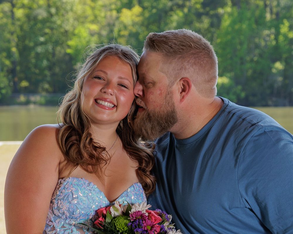 A smiling bride in a floral dress holds a bouquet as a man in blue kisses her cheek outdoors near a lake.