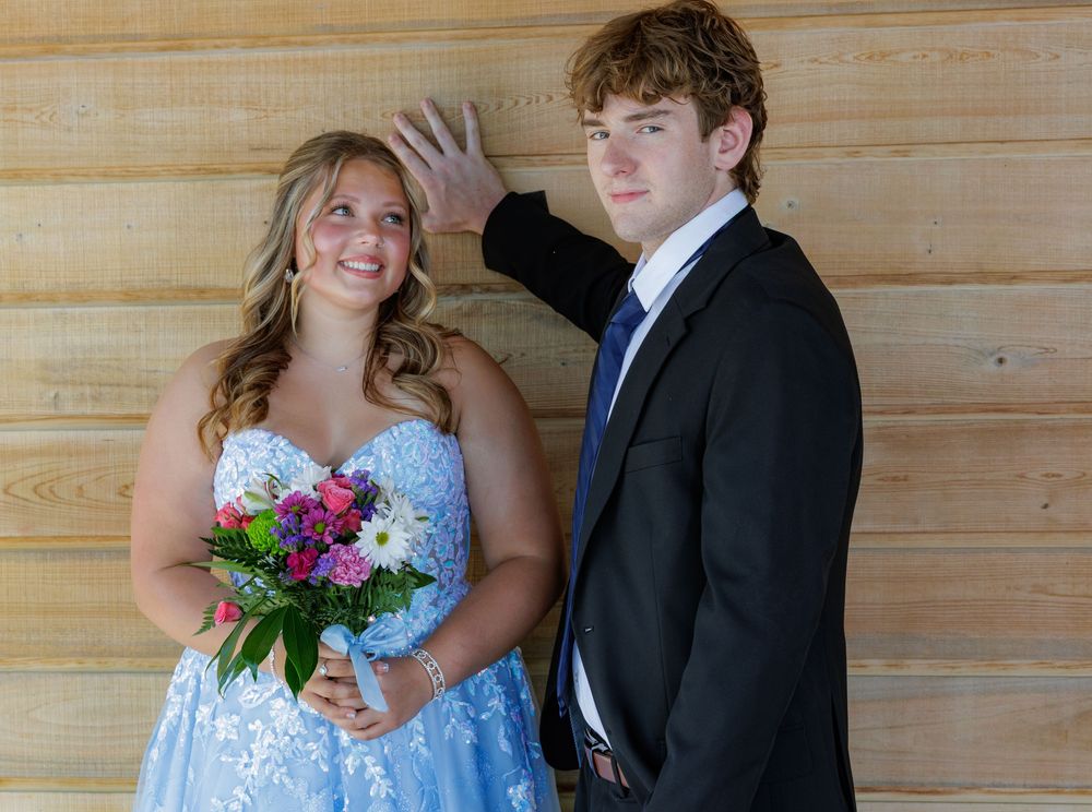 Teen couple in prom attire, girl in blue gown holding bouquet, boy in black suit with blue tie, posing indoors.