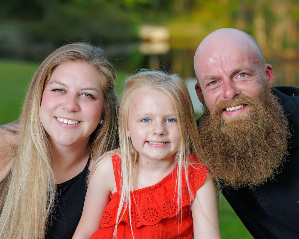 A smiling family of three poses outdoors, featuring a blonde woman, a young girl in red, and a bearded bald man.