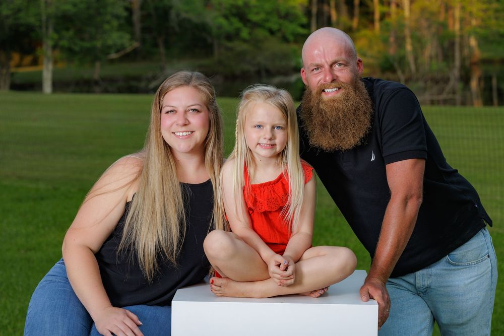 Family portrait of two adults and a young girl in red dress sitting on white cube outdoors on green lawn.