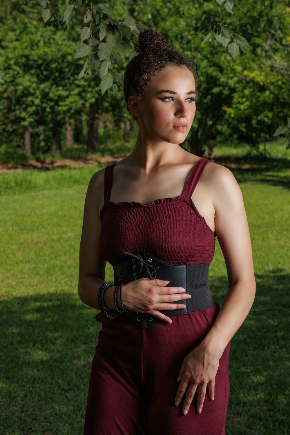 Young woman in burgundy jumpsuit with black corset belt poses outdoors in lush green park setting.
