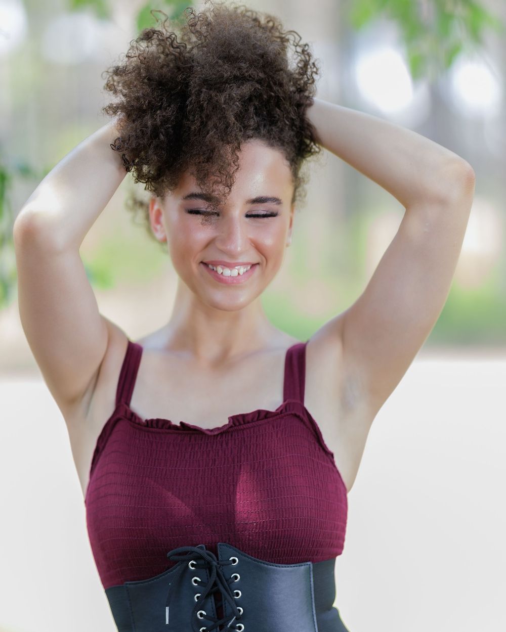 Smiling young woman with curly updo wearing burgundy crop top and black lace-up corset outdoors.