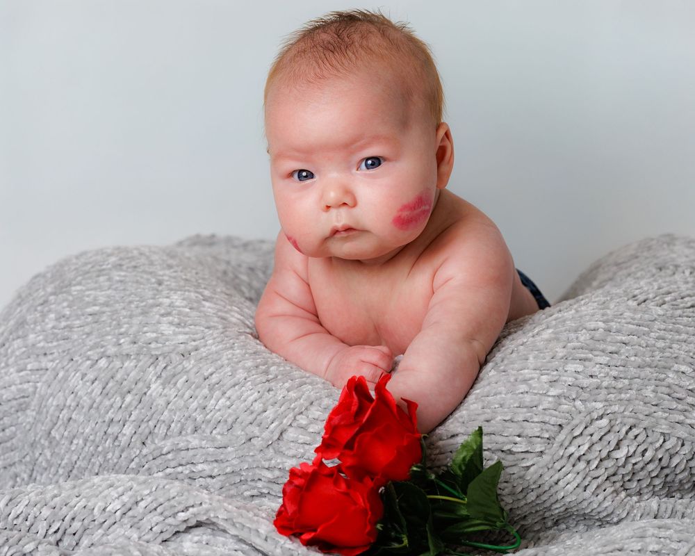 Baby lying on grey blanket with red flower prop in studio setting.