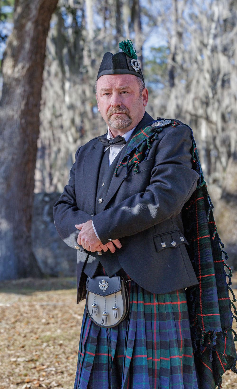 A person wearing traditional Scottish kilt and formal attire poses outdoors against a wooded background.