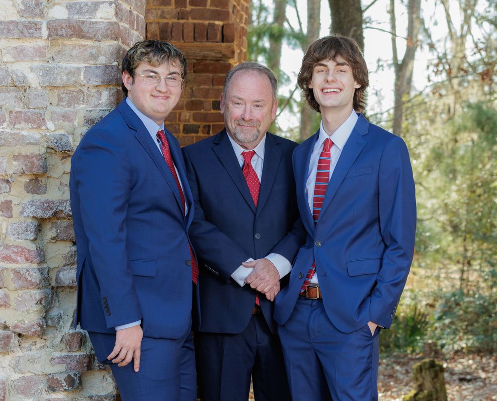 Group of young men in navy suits and red ties pose for formal photo outdoors.