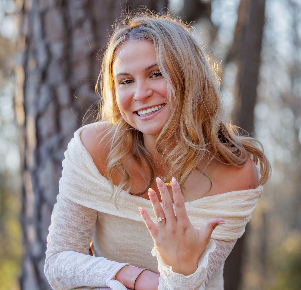 Person in cream colored sweater showing off engagement ring in winter forest setting.