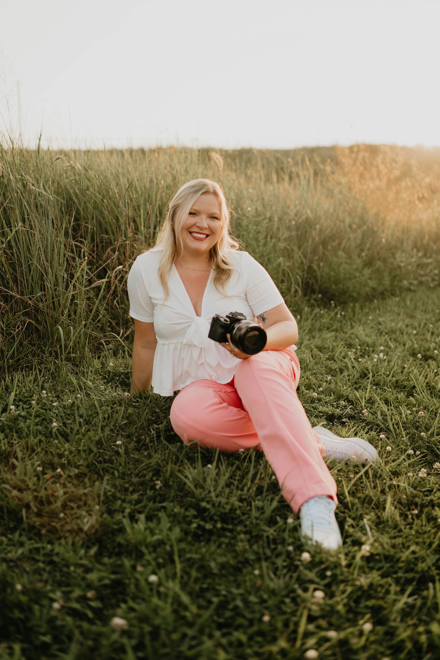 A young woman with blonde hair sitting on the ground outside at sunset. She is holding a camera and smiling, looking very happy.