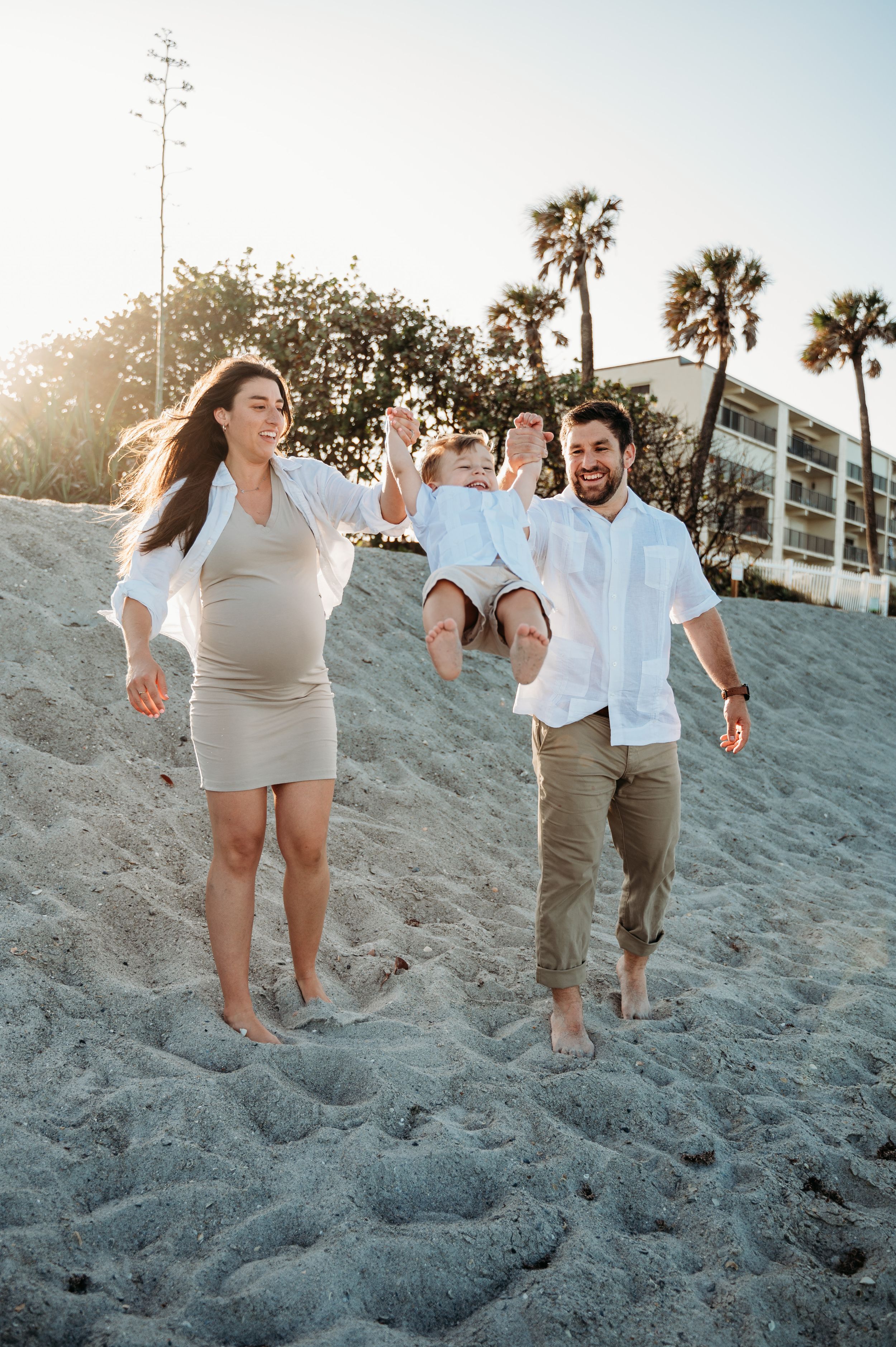 A family enjoys a playful moment together on a sandy beach at sunset with palm trees in the background.