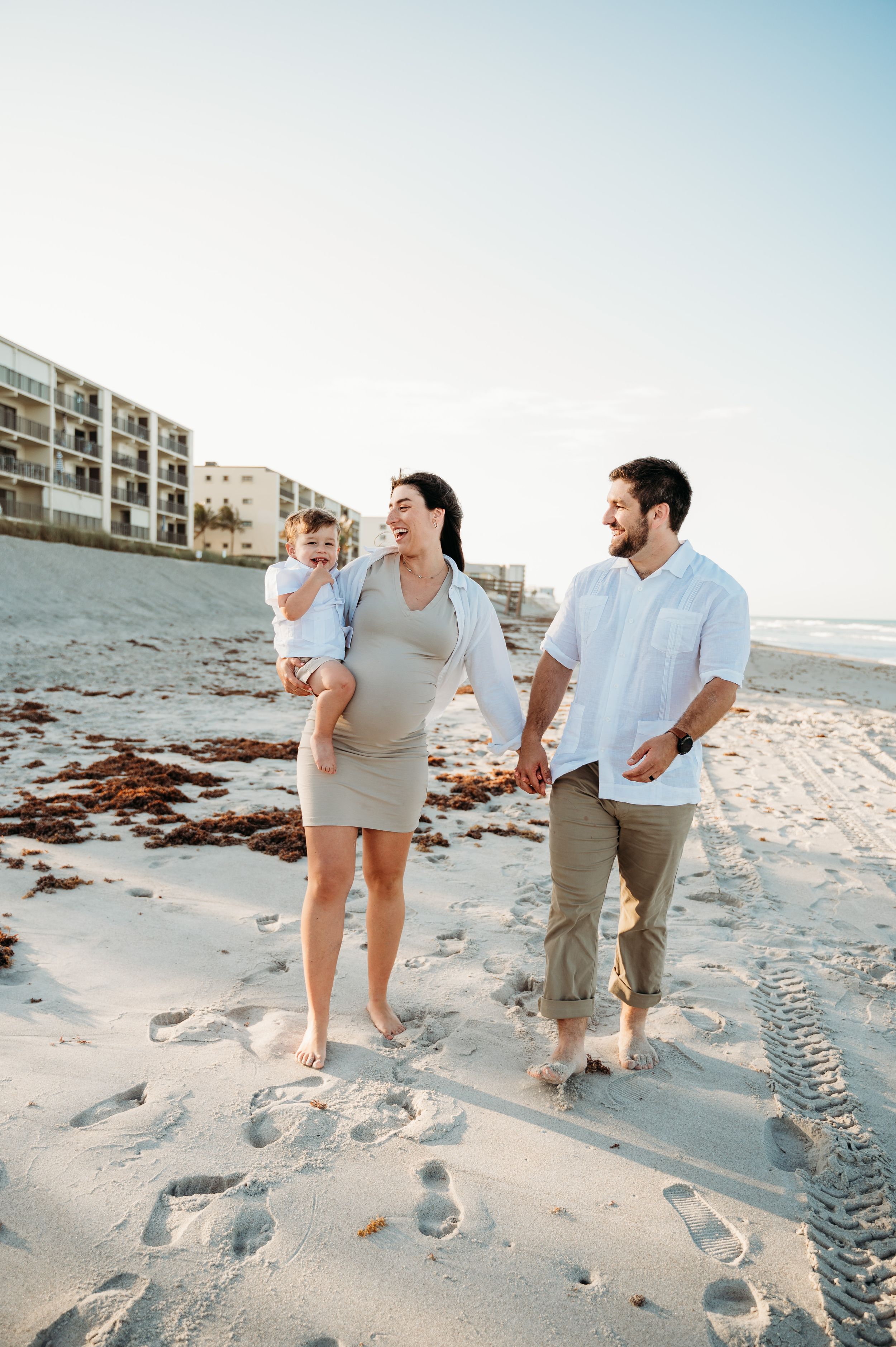 A family walks together along a sandy beach at sunset with apartment buildings in the background.