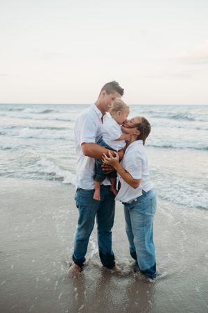 A family shares a tender moment together while standing on a beach at sunset with ocean waves in the background.