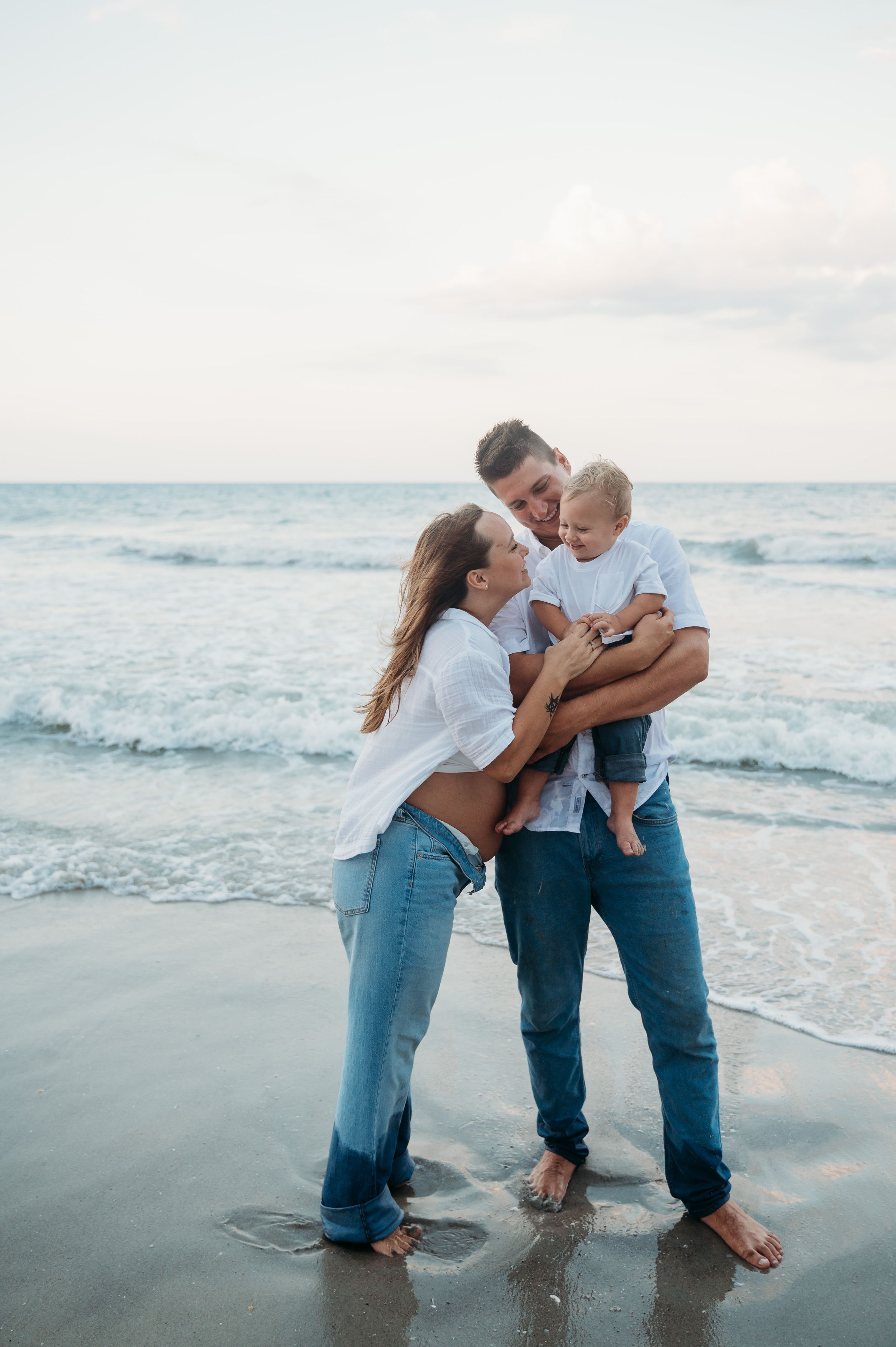 Family shares tender moment together while standing barefoot on wet sandy beach at sunset with ocean waves in background.