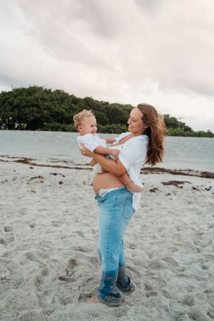 A pregnant woman in jeans and white shirt holds a toddler while standing on a sandy beach with trees in background.