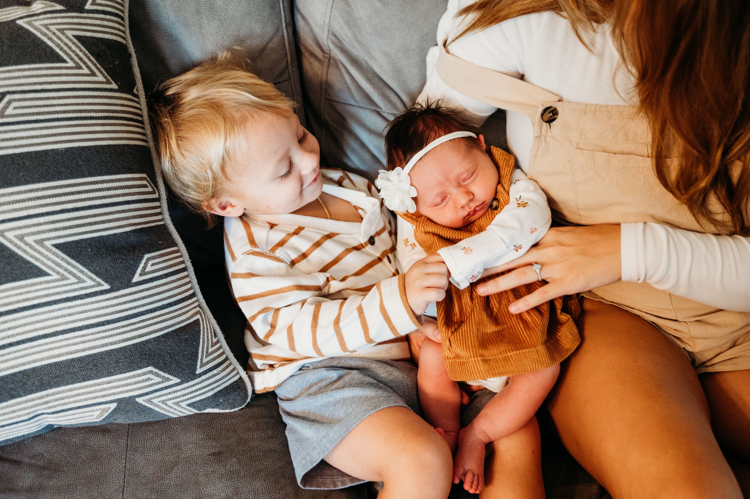 A toddler in a striped shirt sits on a gray couch while meeting a newborn baby being held by someone.