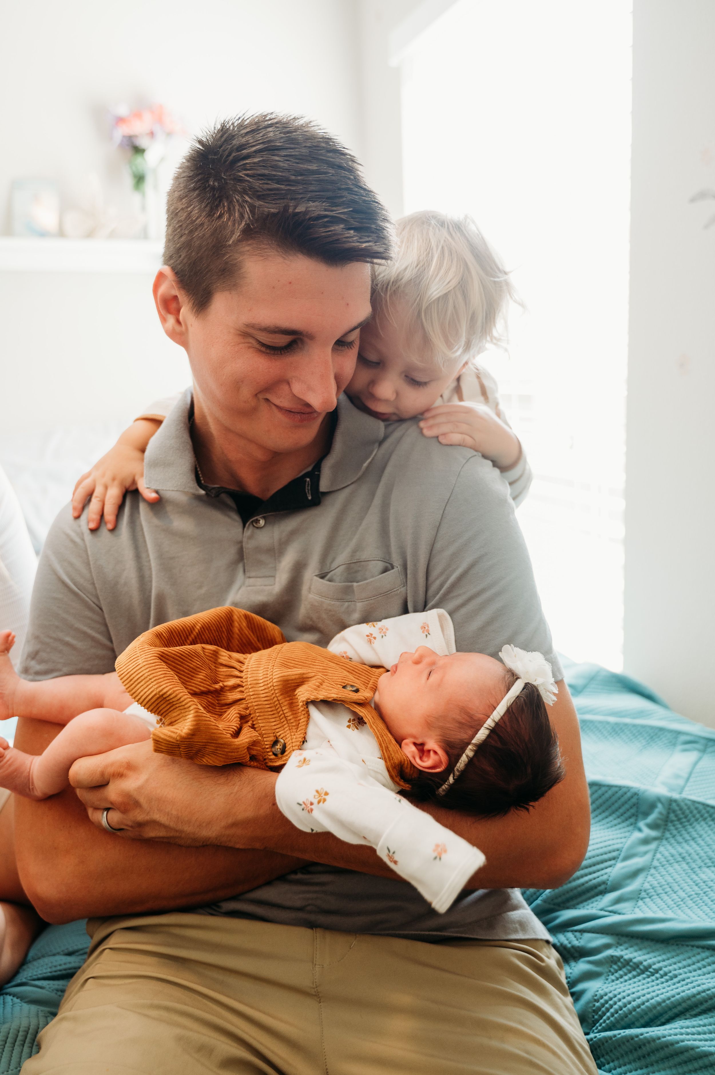A tender family moment shows a parent holding a newborn baby while another child embraces them from behind on a bed.