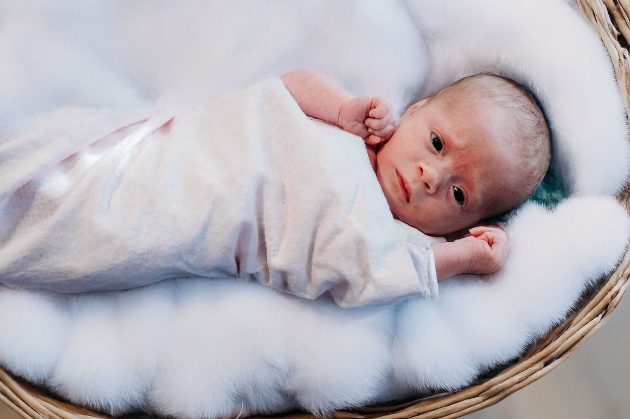A newborn baby wearing white clothing lies on soft white bedding in a woven basket.