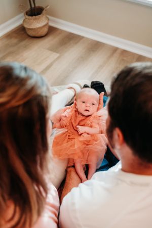A baby wearing a peach-colored dress lies on a white surface while two people look on in a room with hardwood floors.