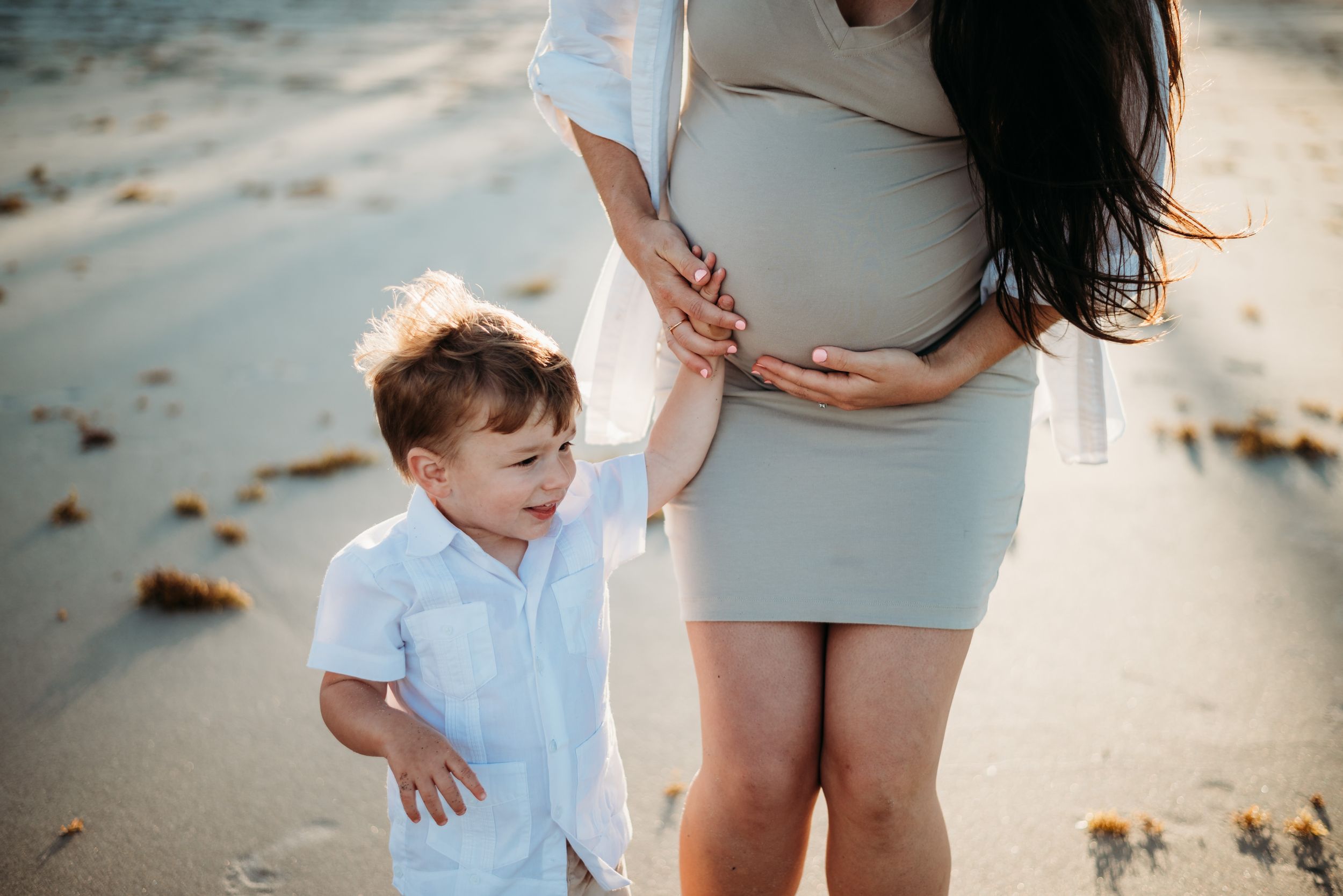 A pregnant woman in a gray dress stands on a beach with a young child in a white shirt.