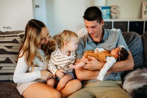 Family sits together on gray couch while toddler in striped shirt meets newborn sibling for first time.