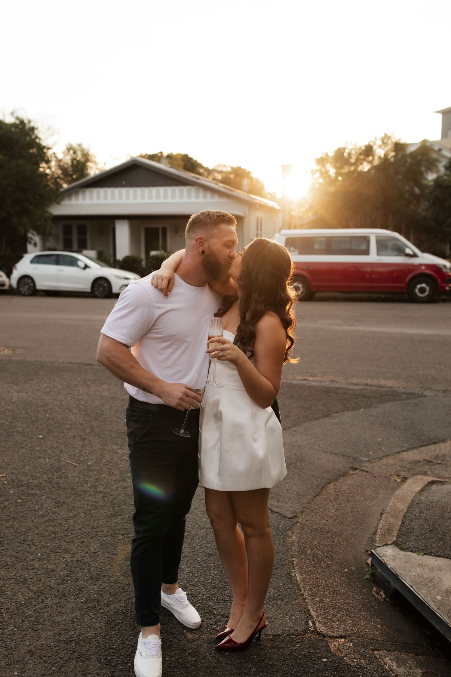 Couple shares a tender moment at sunset in front of a white house with cars in background.