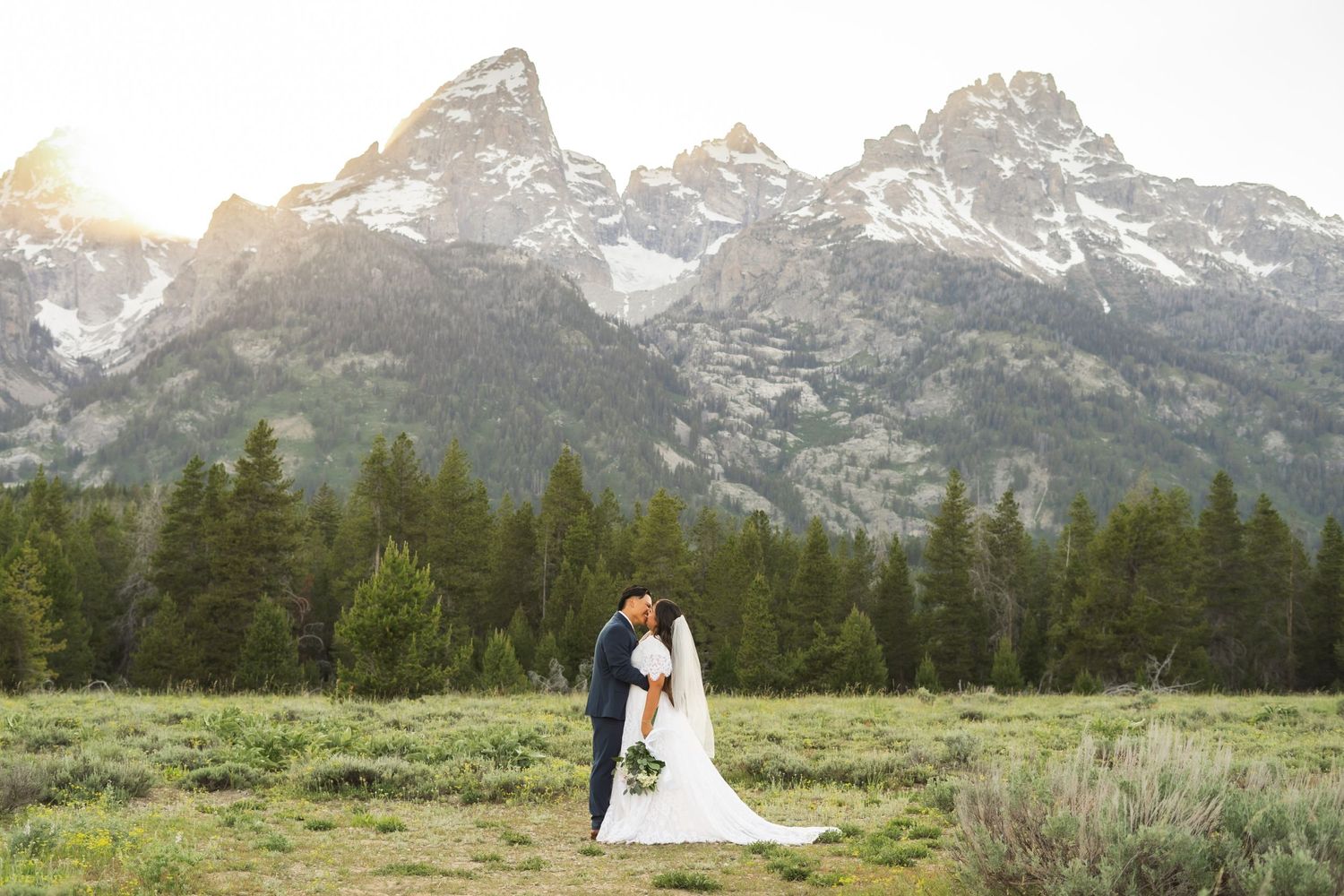 A couple embracing in a meadow with the dramatic Grand Teton mountains rising behind them.
