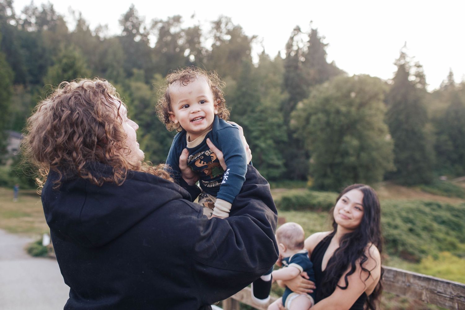 A father spins around his son in a playful way while mom looks on, during a family photo session outdoors in the Seattle area