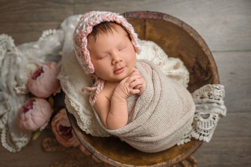 Newborn baby sleeping peacefully in a wooden bowl, wrapped in beige, wearing a pink knit bonnet with floral accents.