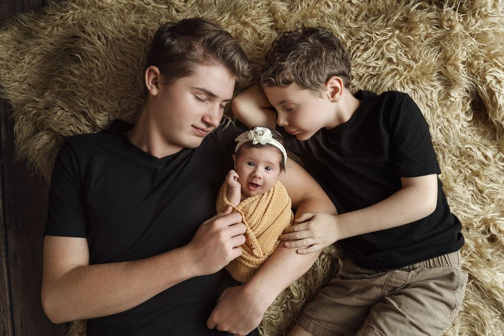 Two brothers lying on a fur rug, tenderly holding a sleeping newborn baby wrapped in a yellow swaddle.