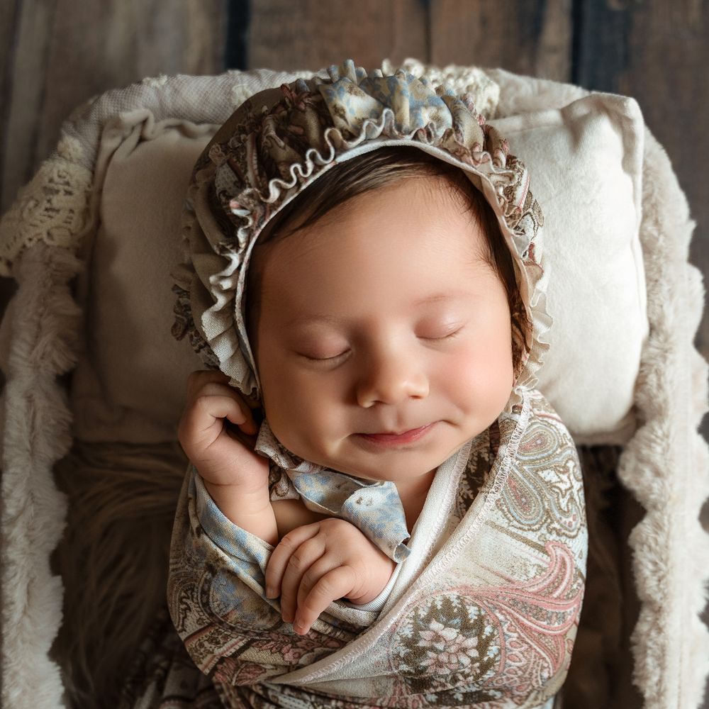 Sleeping newborn baby wrapped in paisley swaddle wearing a vintage lace bonnet, posed in a rustic wooden bowl.