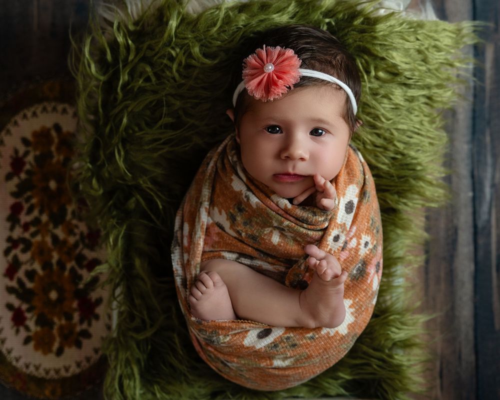 Newborn baby girl wrapped in floral swaddle with pink headband on green fur backdrop.