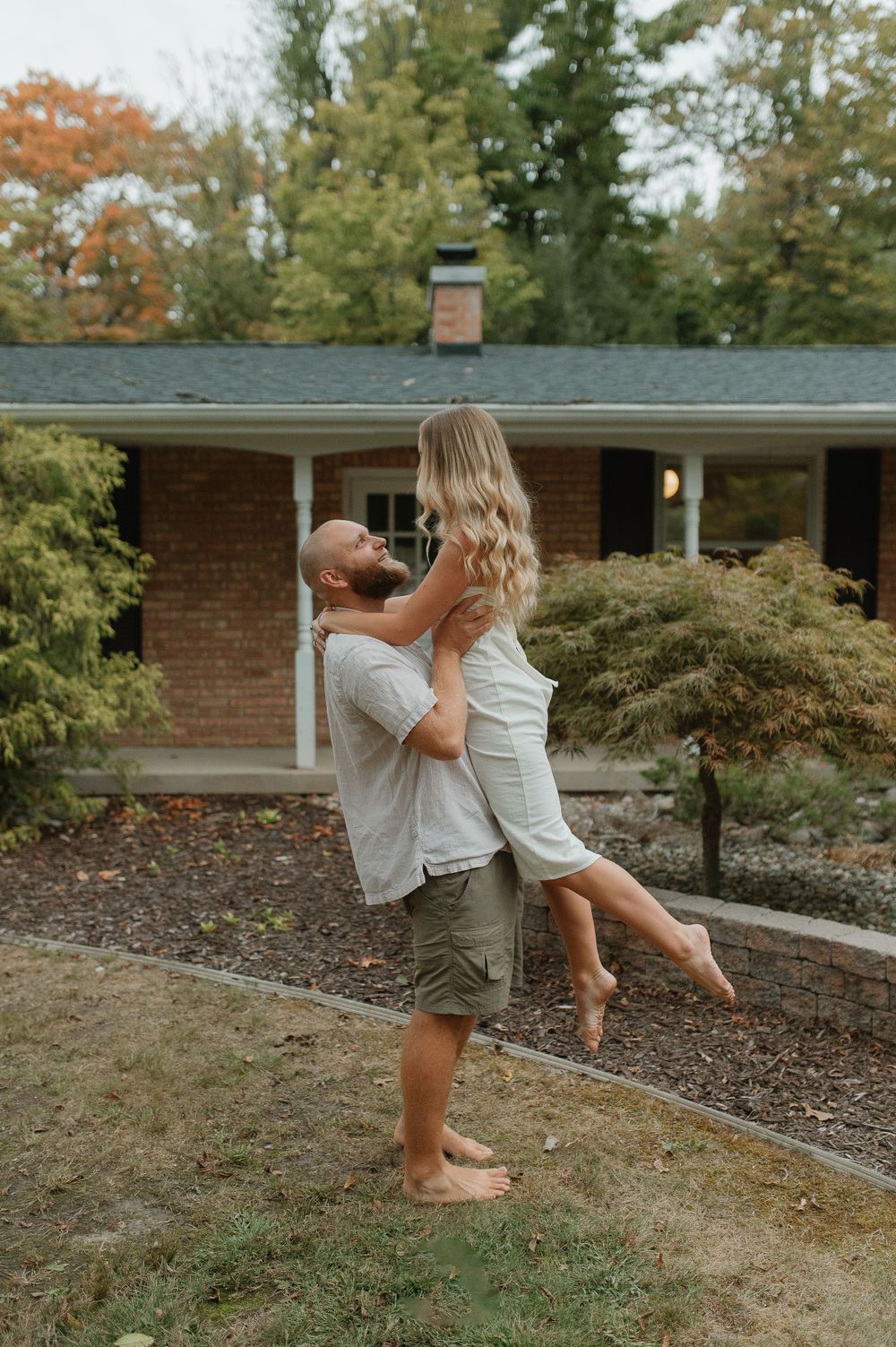 Romantic outdoor moment captured in front of a brick house with autumn trees in the background.
