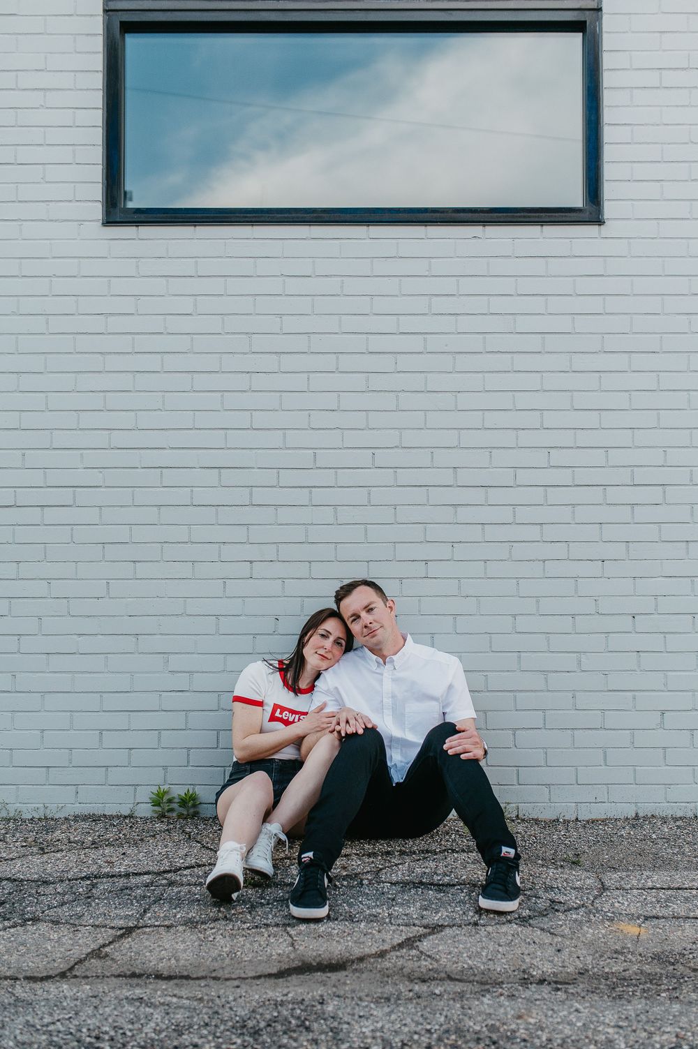 Casual portrait of a couple sitting against a white brick wall beneath a window.