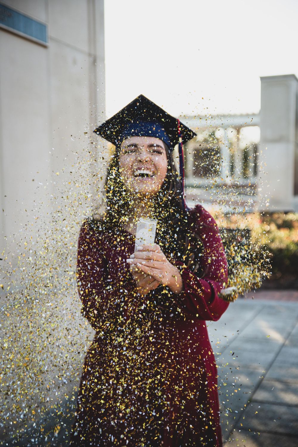 A joyful graduate in a burgundy gown celebrates by popping gold and silver confetti.