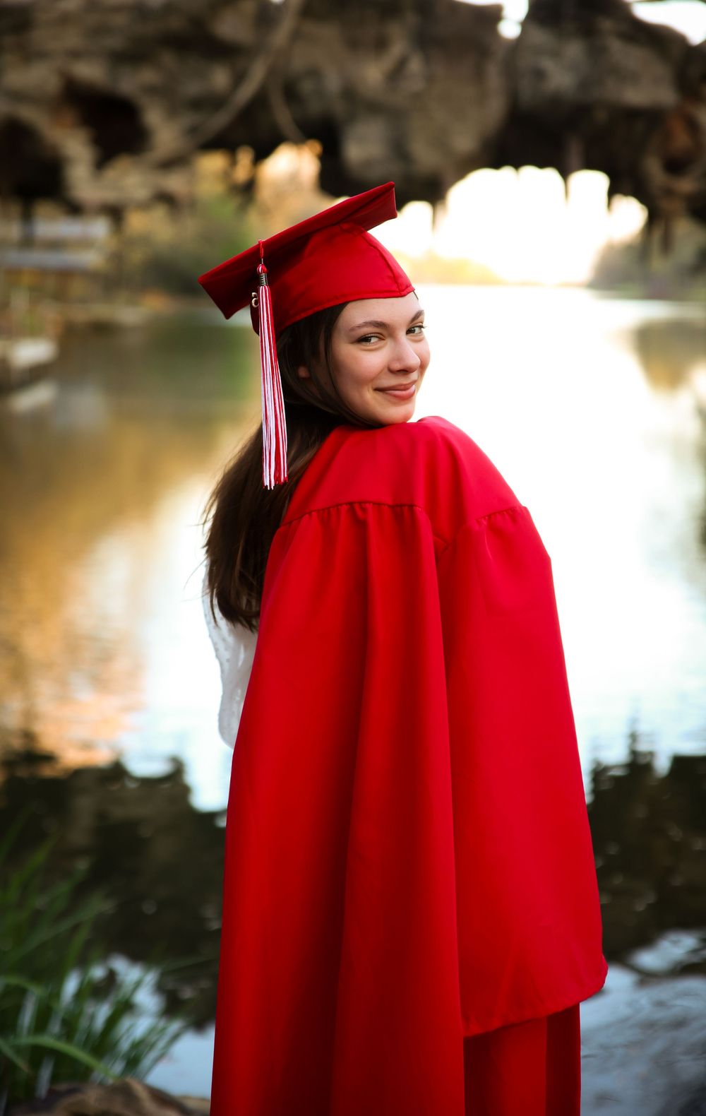 A smiling young woman poses in a red graduation cap and gown near a reflective pond.