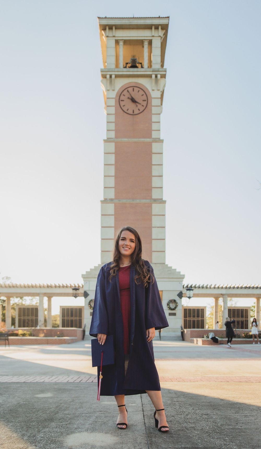A female graduate in a navy gown stands before a tall campus clock tower on a clear day.