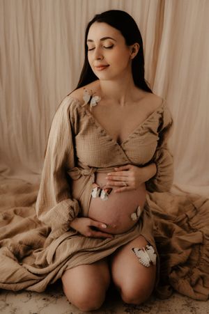 An artistic maternity photo of a woman in beige dress with butterflies against neutral backdrop.