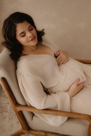 Elegant maternity photo of expectant mother reclining in cream chair wearing white dress.