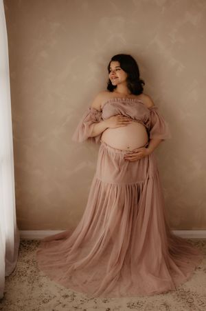 Full-length maternity portrait in blush pink off-shoulder gown against neutral wall.