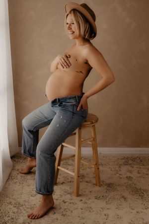 Relaxed maternity portraits featuring blue jeans and wooden chair against neutral wall.