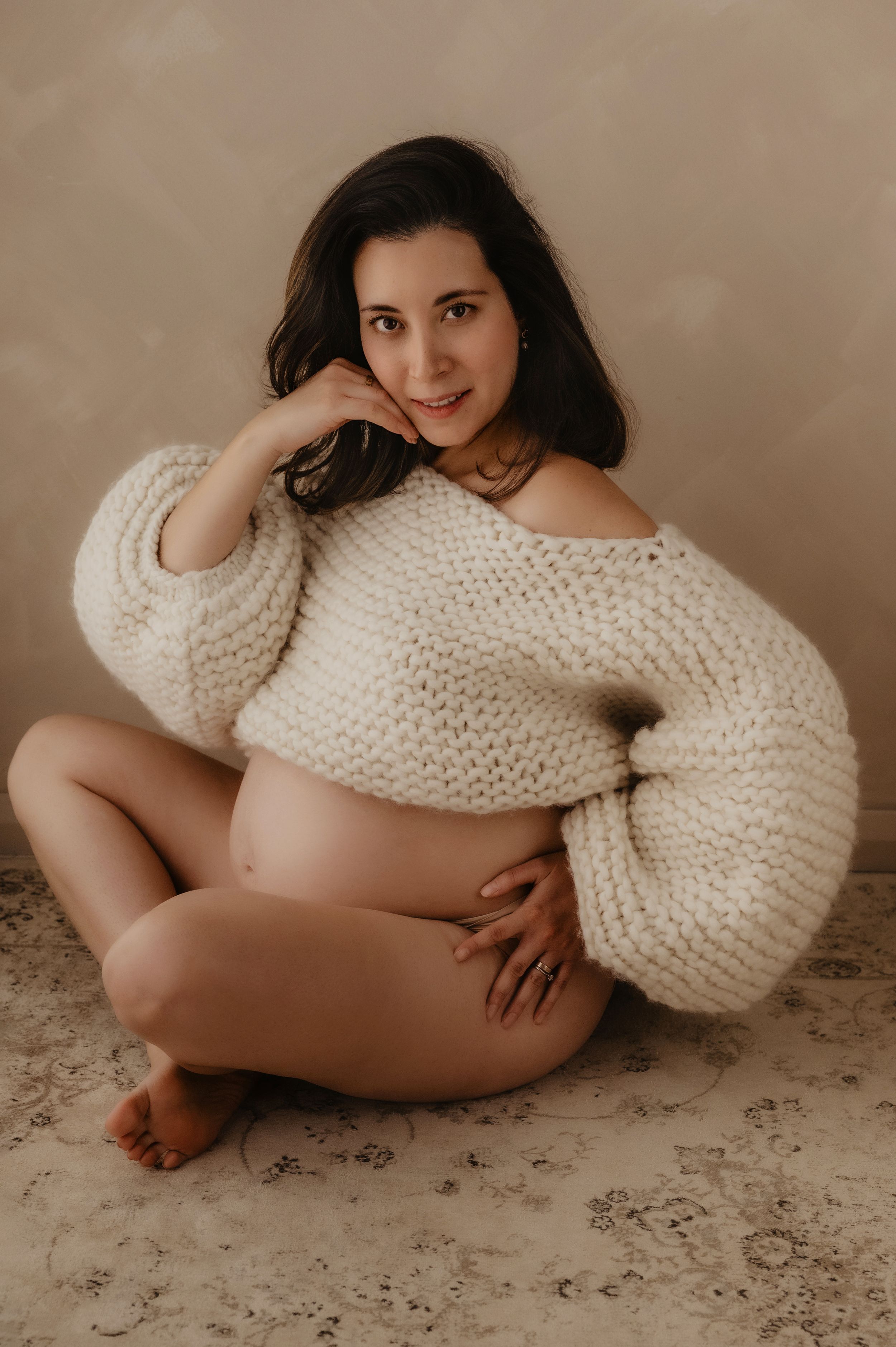 Artistic maternity photoshoot featuring a cozy cream knit sweater against a neutral beige backdrop.