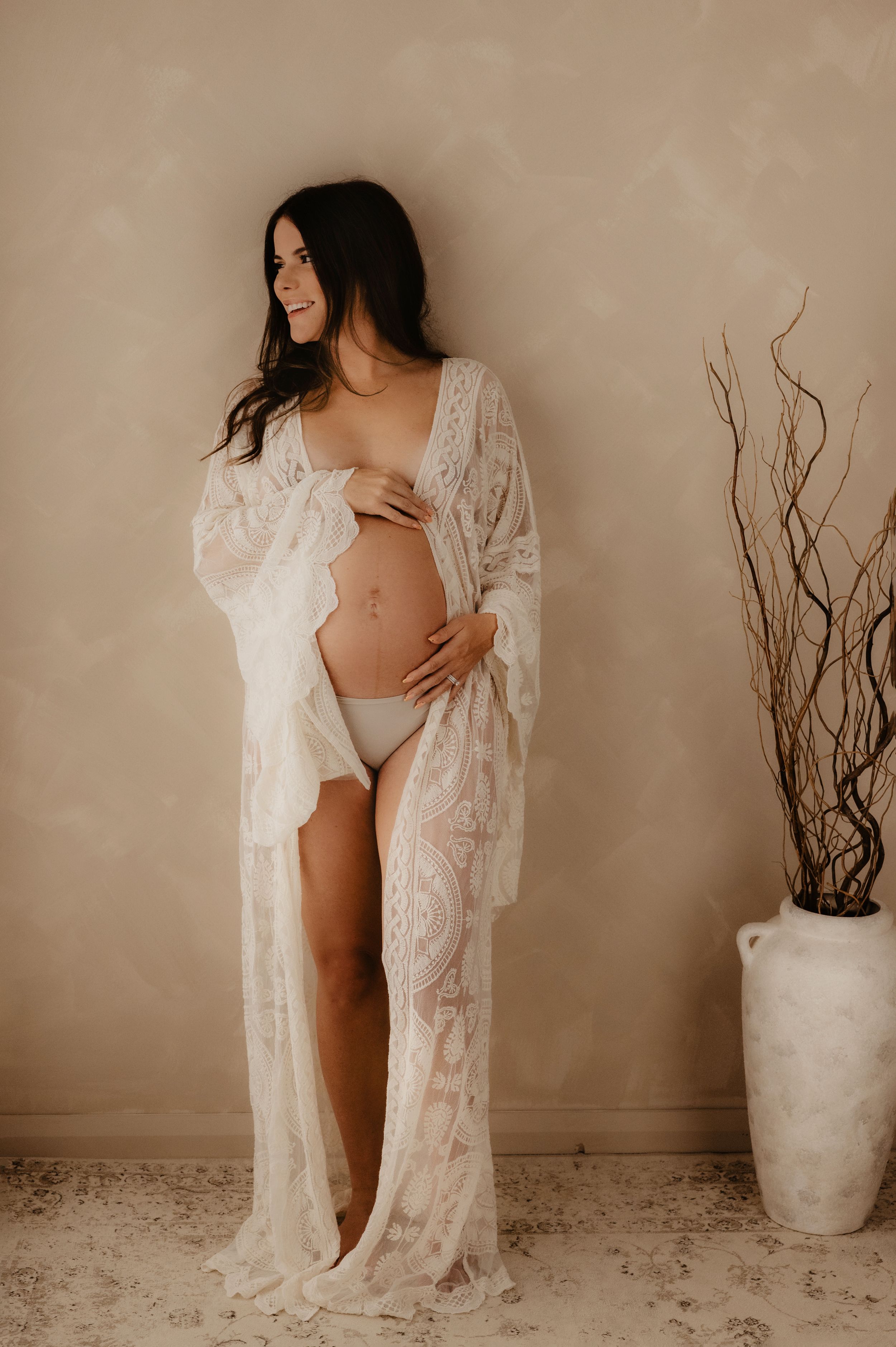 A woman wearing a white lace robe poses for a maternity photo against a neutral beige background with a decorative vase.