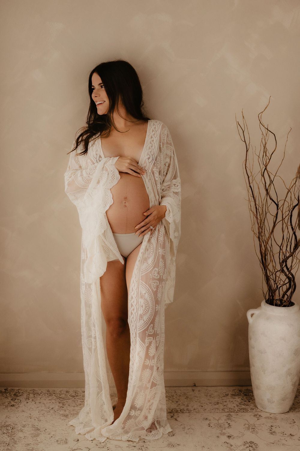 A woman wearing a white lace robe poses for a maternity photo against a neutral beige background with a decorative vase.