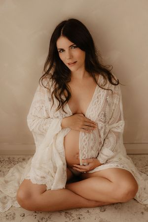 Artistic maternity photo featuring a woman in a white lace robe sitting on floor against beige backdrop.