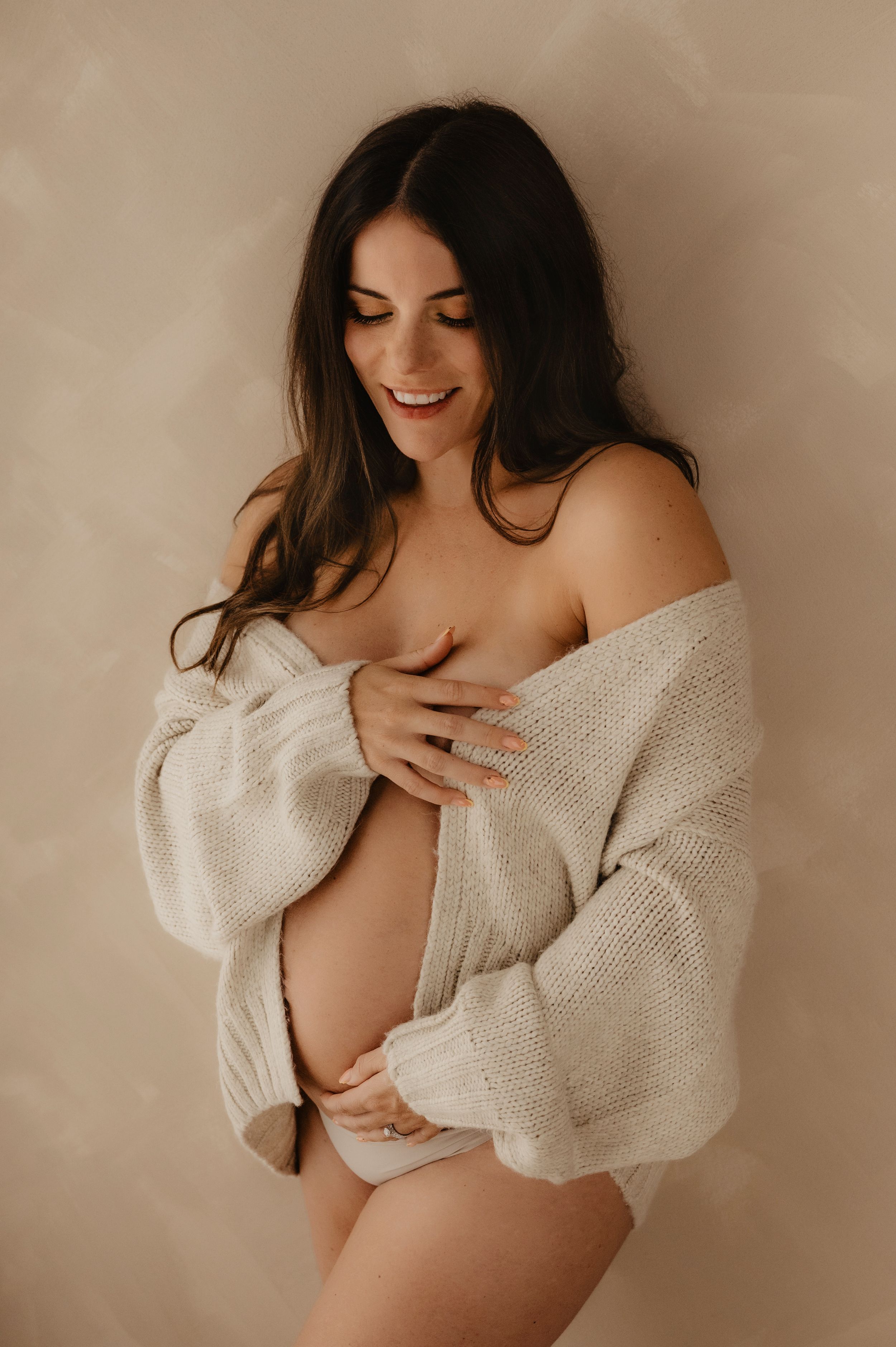 Artistic maternity portrait of an expectant mother in a cozy white sweater against neutral backdrop.